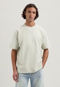 Tricou scurt cu mânecă, de culoare verde deschis, realizat dintr-un material texturat, având un gât rotund și un branding subtil pe față. Purtați cu blugi de culoare albastru deschis.