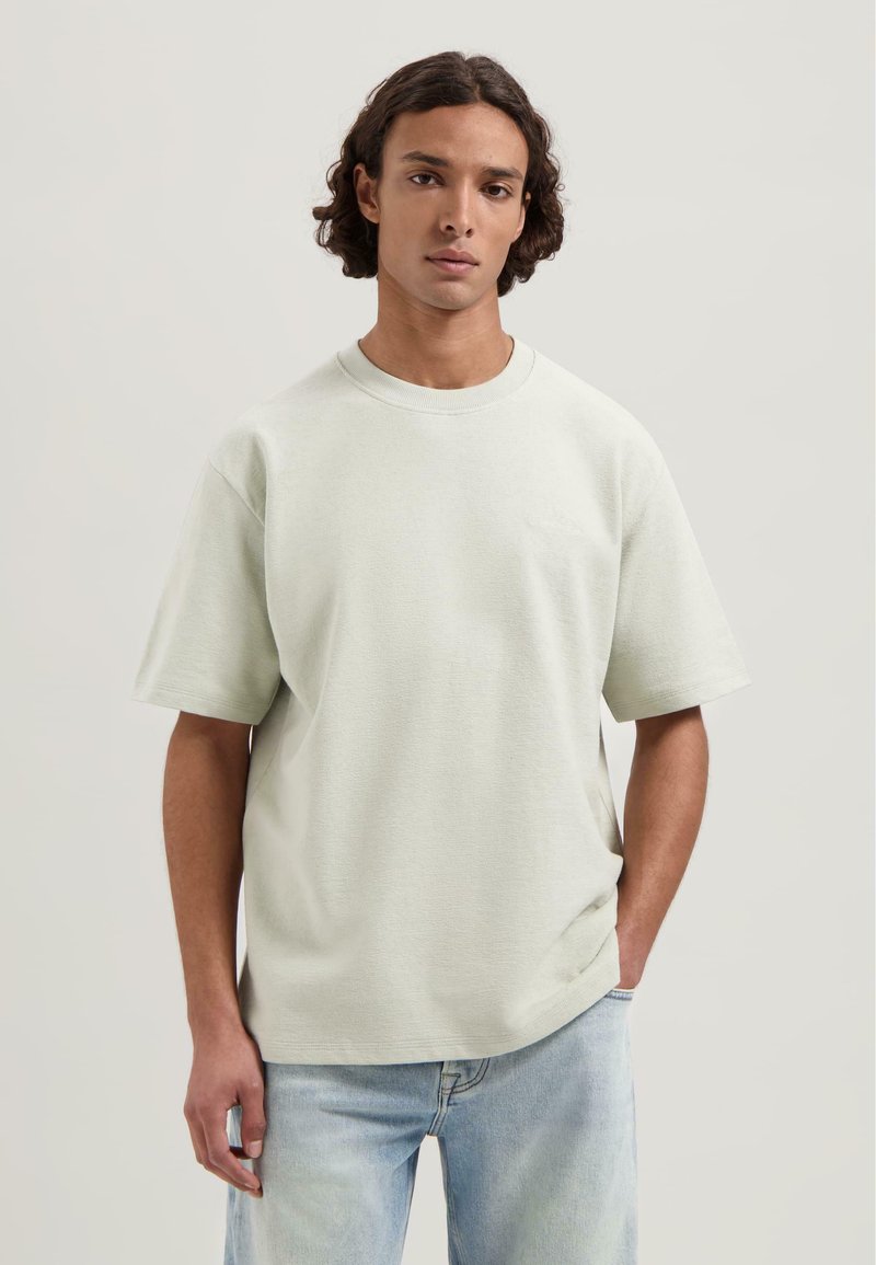 Tricou scurt cu mânecă, de culoare verde deschis, realizat dintr-un material texturat, având un gât rotund și un branding subtil pe față. Purtați cu blugi de culoare albastru deschis.