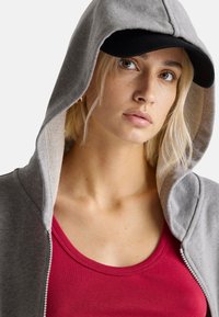 Felpa grigia con zip e cappuccio, abbinata a una canotta rossa. La felpa ha una texture morbida e un leggero sottopelo; una cappellino nero è indossato sopra.