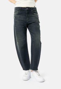 Jean en denim bleu foncé avec une coupe décontractée, des coutures bien visibles et de petits détails usés. Associé à des baskets blanches.