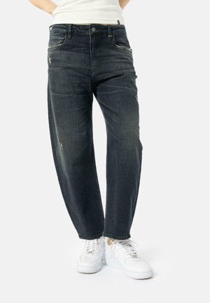 Donkerblauwe denim jeans met een relaxte pasvorm, zichtbare stiksels en kleine versleten details. Gecombineerd met witte sneakers.