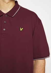 Homme portant un polo bordeaux avec un logo d'oiseau jaune sur la poitrine et une bordure gris clair au col et aux manches.