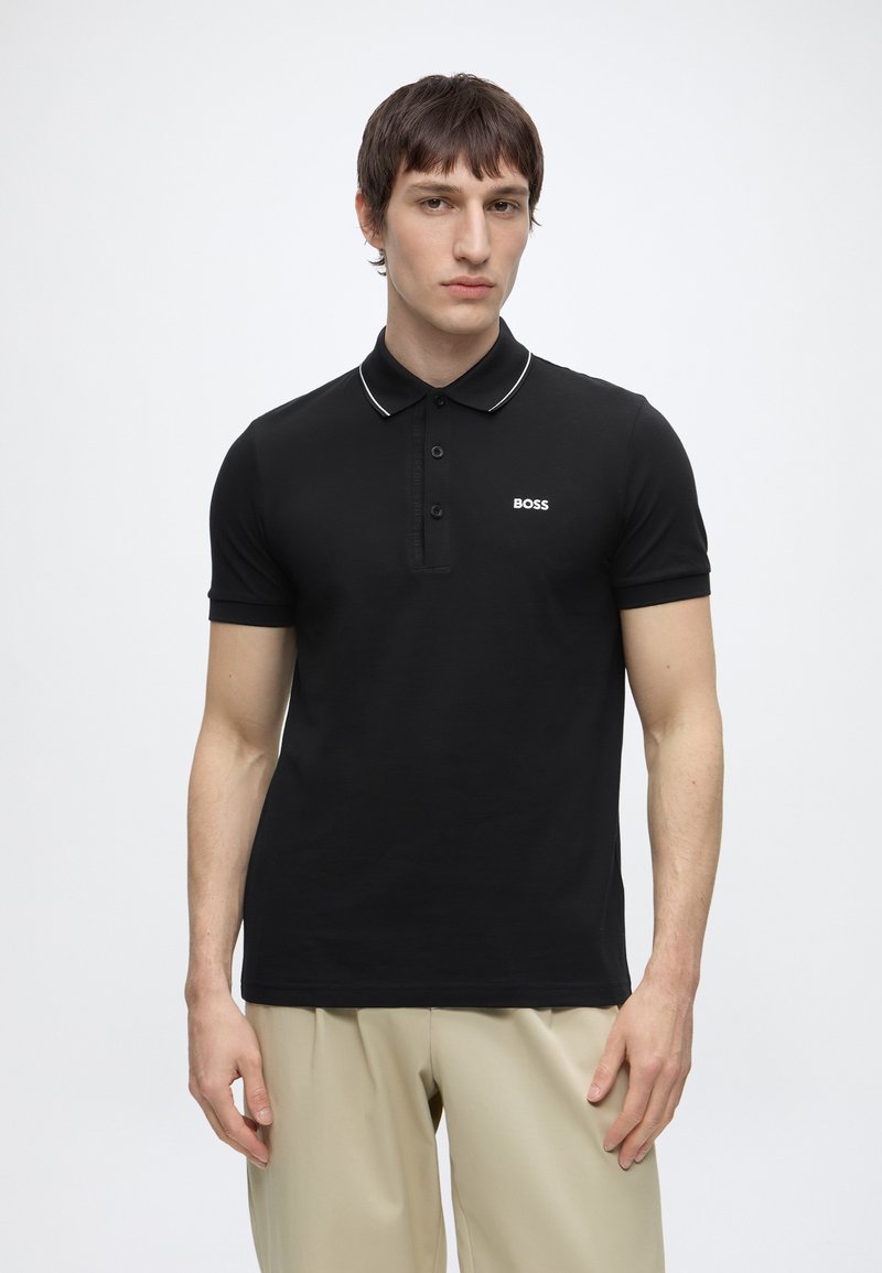 Mand iført sort kortærmet poloshirt med hvide detaljer og "BOSS"-logo, kombineret med beige bukser, stående mod en hvid baggrund.