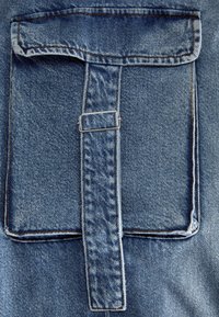 Denimska žep s poklopcem, ki vključuje kovinsko zanko in kontrastno šivanje. Tkanina je srednje modra z teksturirano površino.