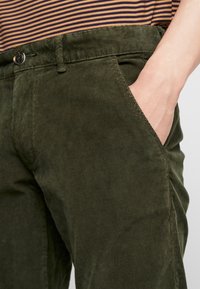 Spodnie ze sztruksu w kolorze oliwkowej zieleni o prostym kroju, z bocznymi kieszeniami i zapięciem na guzik w pasie. Subtelna prążkowana faktura.