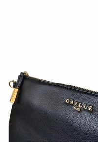 Clutch in pelle nera con superficie testurizzata, accessori in oro, chiusura con zip superiore e logo inciso "GAËLLE PARIS" sul fronte.