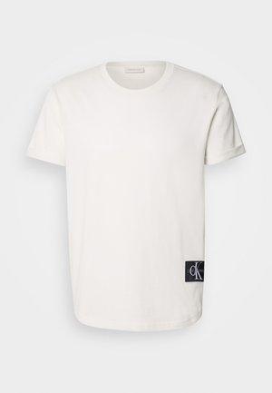 Valkoinen puuvillapaita, jossa on pyöreä kaula-aukko ja lyhyet hihat, ja jonka alaosassa vasemmalla puolella on musta logopatch. Sileä pinta ja suora leikkaus.