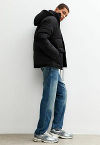 Chaqueta acolchada negra con capucha, tela texturizada, dos bolsillos frontales, combinada con jeans de color azul claro y zapatillas gris. Ajuste casual.