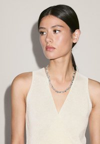 Collana a stringa con perle di argento arrotondate, indossata con un top senza maniche di un leggero beige. Si notano la textura a maglia e il design a scollo a V.