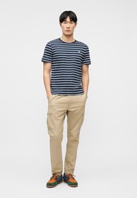 Νέος άνδρας στέκεται με τα χέρια στις τσέπες, φοράει μπλε ναυτικό και λευκό ριγέ μπλουζάκι, μπεζ παντελόνι καμπαρντίνα και πολύχρωμα δερμάτινα παπούτσια με κορδόνια σε λευκό φόντο.