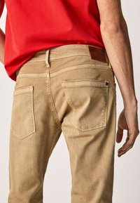 Jeans de mezclilla beige con una textura suave, que cuentan con cinco bolsillos, detalles de costura sutiles y una etiqueta de marca de cuero marrón en la cinturilla.