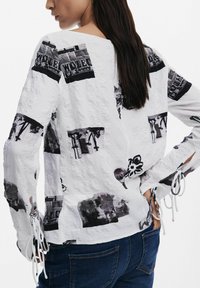 Bluză albă cu mâneci lungi, prezentând imprimeuri grafice negre, țesătură texturată, manșete deschise cu șnururi și un croi lejer.