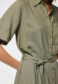 Abito verde oliva a bottoni realizzato in tessuto texturizzato, caratterizzato da maniche corte, una tasca frontale e una cintura legata in vita.