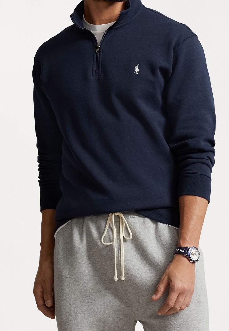 Mørkeblå quarter-zip sweatshirt med ribbede manchetter og krave, med et lille hvidt logo på venstre bryst. Bæres over grå træningsbukser.