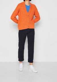 Oranssi neulecardigan, jossa on V-pääntie, ribatut hihansuut ja viisi nappia. Päälläsinisen paidan kanssa, tummansiniset caprimittaiset housut ja valkoiset sneakerit.