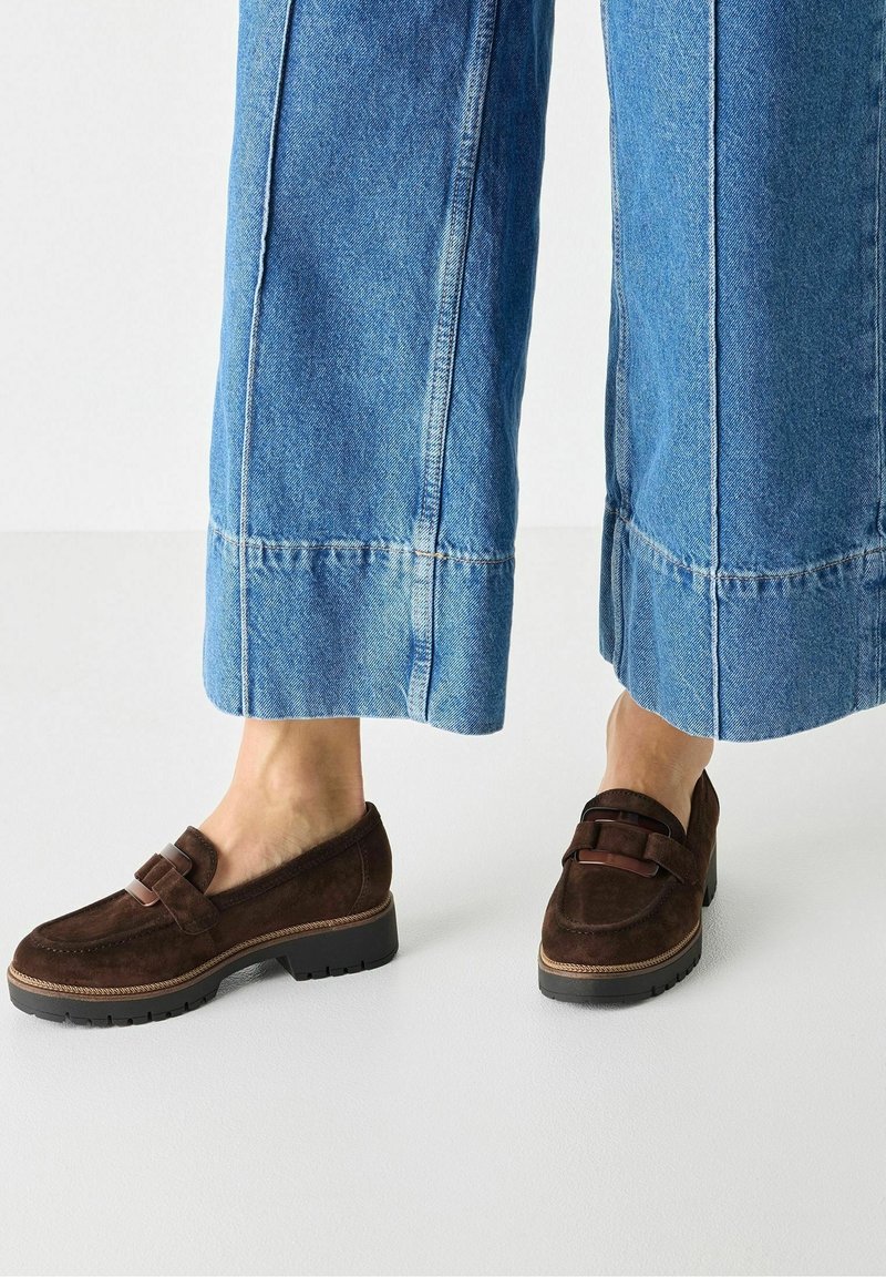 Personne portant un jean bleu à jambes larges et des mocassins en daim marron avec des semelles noires épaisses, debout sur un sol blanc.