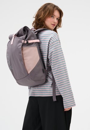 Junge Frau trägt ein gestreiftes Langarmshirt und schwarze Hose, hat einen großen grauen und pinken Rucksack über einer Schulter.