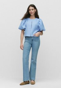 Ljust blå puffärmad topp i texturerat tyg, med framknäppning, tillsammans med ljusblå utsvängda jeans och beige lägenheter. Enkelt bakgrund.