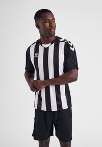 Sort-hvidstribet trøje med korte ærmer, featuring en V-hals og sporty tekstur. Båret med sorte shorts og et humel-logo på brystet.