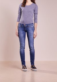 Camiseta de manga larga gris, vaqueros ajustados azules y zapatos tipo slip-on estampados con suela blanca. Textura lisa; diseño casual.