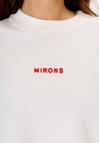 Weißer Sweatshirt mit roten gestickten Text, der "MIRONS" liest. Es hat einen klassischen Rundhalsausschnitt und eine weiche, glatte Stoffbeschaffenheit.