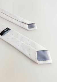 Corbata blanca texturizada con pequeños puntos beige, forro gris visible en ambos extremos y una etiqueta negra de la marca Next en el reverso.