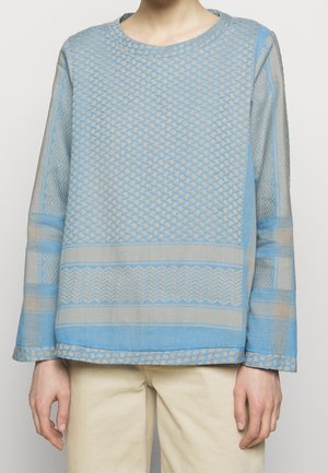 Person trägt ein hellblau gemustertes Langarmshirt mit beige Zickzack- und Punktmustern, kombiniert mit hellbeigen Hosen.