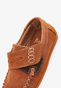Chaussure en cuir marron avec une finition texturée, dotée d'une fermeture à sangle et de détails cousus. Semelle en caoutchouc visible, suggérant un design décontracté.