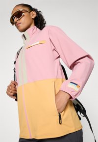 Persona vestida con una chaqueta de color bloque en rosa y amarillo, con bolsillos con cremallera, franja reflectante, gafas de sol y llevando una mochila negra.