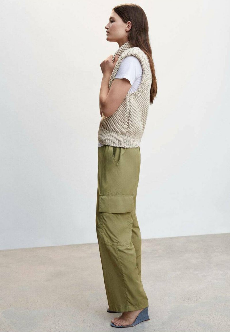 Beżowy, bezrękawnik z dzianiny noszony na białej koszulce, w połączeniu z luźnymi oliwkowymi spodniami cargo i szarymi sandałami na koturnie.