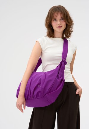 Junge Frau, die ein weißes Oberteil und schwarze Hosen trägt, trägt eine große lila Umhängetasche mit breitem Riemen quer über ihren Körper.