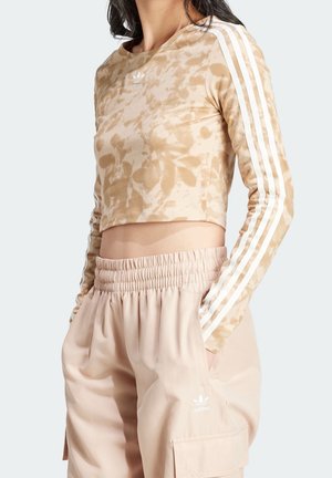 Kobieta ubrana w beżowy, długorękawowy krótki top tie-dye z białymi paskami oraz beżowe spodnie z elastycznym pasem i kieszeniami, obie rzeczy marki Adidas.
