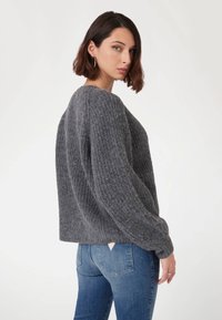 Grauer, strukturierter Pullover mit lockerer Passform, geripptem Muster und weiten Ärmeln, kombiniert mit blauen Jeans, die ein kleines, dreieckiges Etikett haben.