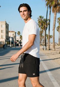 Mlad moški v beli majici in črnih kratkih hlačah hodi po sončni sprehajalni poti ob plaži s palmami in stavbami v ozadju.