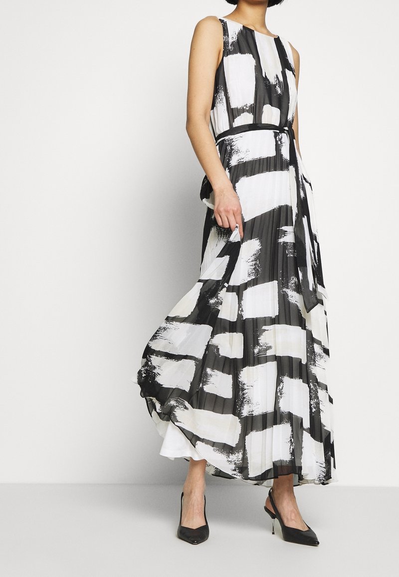 Femme portant une robe maxi plissée sans manches à motif abstrait noir et blanc avec des talons hauts noirs, debout sur un fond blanc.