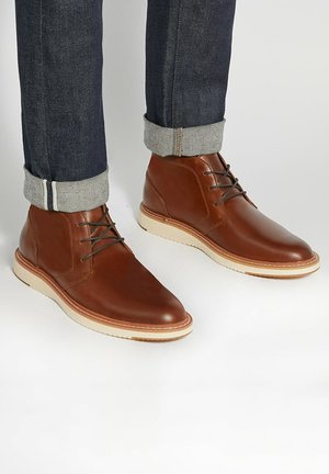 Brown leather ankle boots with smooth surface, round toes, and grey laces. Features a cream-colored rubber sole and rolled denim cuffs.