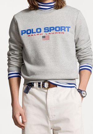Persona vistiendo sudadera deportiva Puma gris con logo de la bandera estadounidense, cuello alto a rayas azules y blancas y puños, pantalones blancos, cinturón a rayas y reloj.
