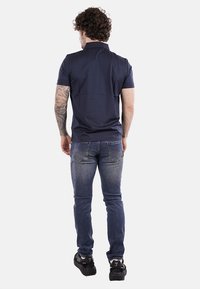 Polo shirt della marina a maniche corte, slim fit, abbinato a jeans blu aderenti e sneakers nere. Tatuaggio visibile su un braccio.