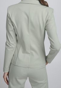 Jasnozielona, dopasowana marynarka z wciętym kołnierzem, gładką fakturą i krawiectwem na miarę, zestawiona z pasującymi spodniami widocznymi na wysokości talii.