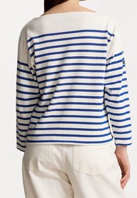 Kvinna som bär en långärmad vit skjorta med blå horisontella ränder och off-white byxor, sedd bakifrån mot en enfärgad bakgrund.