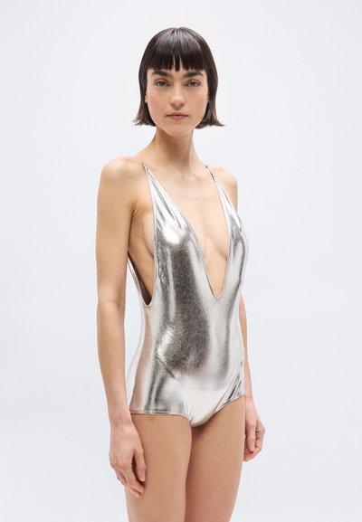 Γυναίκα με κοντά σκουρόχρωμα μαλλιά, φορώντας μεταλλικό ασημί, αμάνικο κορμάκι με βαθύ V ντεκολτέ, στέκεται μπροστά σε ένα απλό λευκό φόντο.