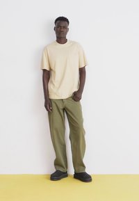 Beige short-sleeve t-shirt with pocket, paired with loose olive green trousers and black shoes, standing on a yellow floor against a white wall.