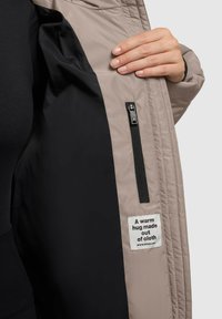 Bēša iešūta jakas iekšpuse ar melnu oderi un sānu rāvējlēdzēja kabatu. Auduma uzlīme lasa: "Silts apskāviens, kas izgatavots no auduma."