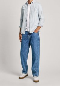 Jasnoniebieska koszula w białe paski z guzikami noszona na białym T-shircie, zestawiona z luźnymi niebieskimi dżinsami i białymi sneakersami.