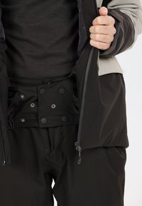 Veste extérieure noire avec des accents gris contrastants, dotée de fermetures à boutons-pression et de poches zippées. Portée par-dessus une couche de base sombre et texturée.