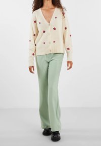 Krämfärgad kofta med rosa blommig broderi, V-ringning och knappar, kombinerad med högmidjade, ljusgröna byxor och svarta platåskor.