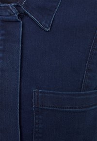 Casaco de ganga azul escuro com colarinho, um bolso no peito e costuras visíveis. A textura é suave com um leve brilho.