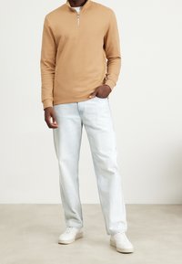 Pullover marrone chiaro con zip a quarto in tessuto testurizzato, abbinato a jeans azzurri sbiaditi e sneakers bianche. Design semplice e casual.