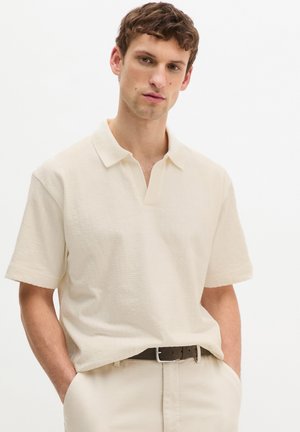 Mann trägt ein cremefarbenes Kurzarmhemd mit Kragen, in beige Hose mit braunem Gürtel gesteckt, Hände in den Taschen, neutraler Hintergrund.