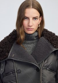 Kobieta z prostymi brązowymi włosami, ubrana w ciemnoszary płaszcz ze sztucznego futra, na szary sweter z golfem, z neutralnym wyrazem twarzy.